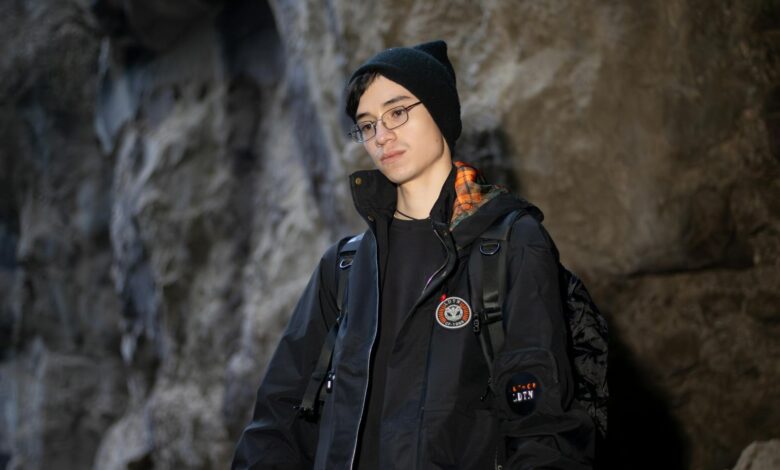 A young adult in a black jacket and beanie inside a rocky cave.