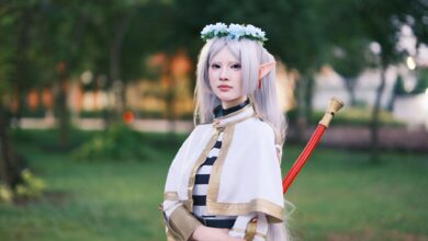 A cosplayer dressed as an elf with long silver hair poses outdoors in a tranquil park.