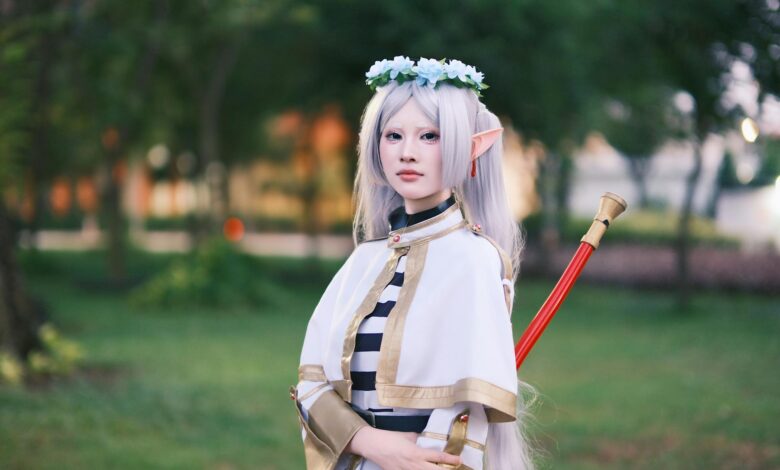 A cosplayer dressed as an elf with long silver hair poses outdoors in a tranquil park.