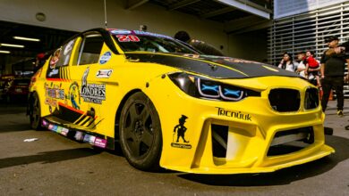 Close-up of a yellow race car with custom decals parked in a garage, surrounded by people.