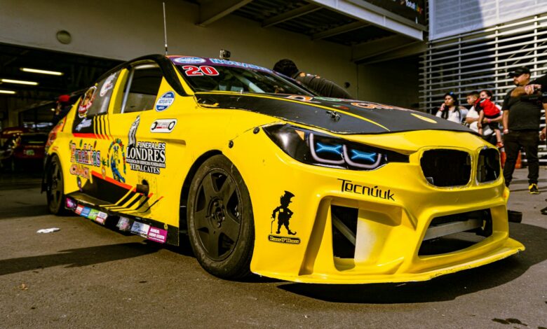 Close-up of a yellow race car with custom decals parked in a garage, surrounded by people.
