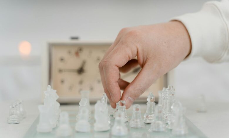 A detailed shot of a hand moving glass chess pieces on a board with a clock in the background.