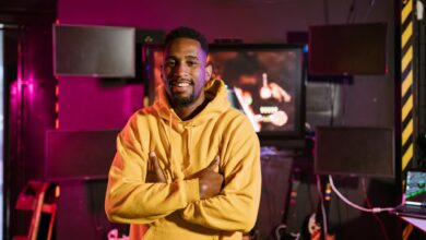 Smiling gamer in a yellow hoodie stands confidently in an e-sports studio with vibrant lighting.