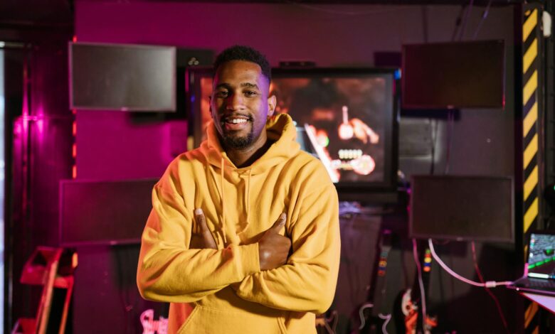 Smiling gamer in a yellow hoodie stands confidently in an e-sports studio with vibrant lighting.