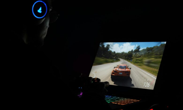 A gamer using a laptop and headphones to play a car racing game in a dark setting.