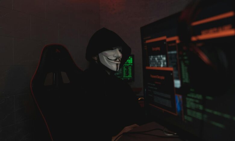 An anonymous hacker wearing a mask working on a computer in a dark room.