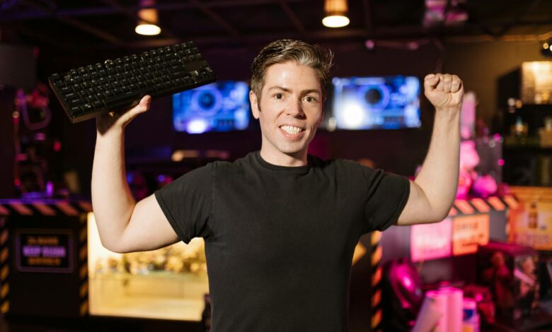 Smiling gamer in black shirt holding keyboard, celebrating indoors with gaming setup.
