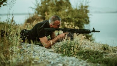 A military sniper in camouflage surveying the landscape with a rifle outdoors near vegetation.