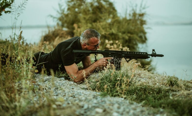 A military sniper in camouflage surveying the landscape with a rifle outdoors near vegetation.