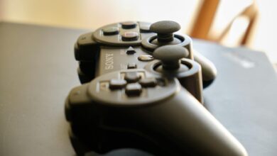 A black wireless game controller with joysticks on a dark surface in soft natural light.