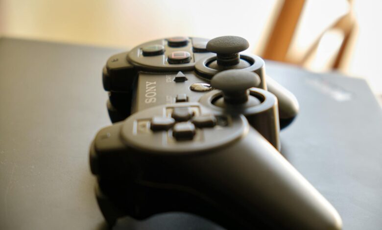 A black wireless game controller with joysticks on a dark surface in soft natural light.
