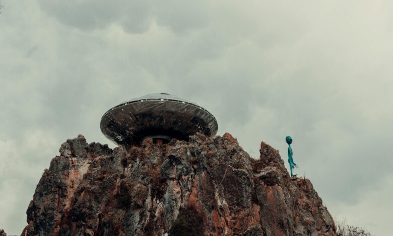 A surreal scene with a UFO and alien standing atop a rugged rock formation against a cloudy sky.