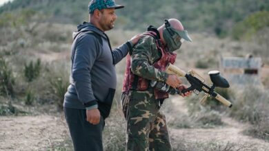 Two men playing paintball outdoors, one wearing camouflage and a mask, in a rural setting.
