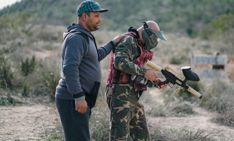 Two men playing paintball outdoors, one wearing camouflage and a mask, in a rural setting.