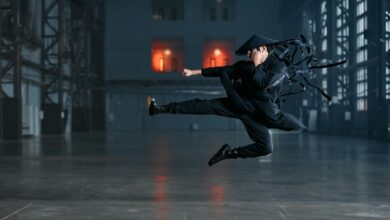 A martial artist performs a jump kick in an industrial setting, wearing a black kimono and conical hat.