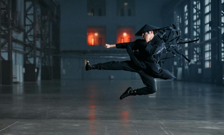 A martial artist performs a jump kick in an industrial setting, wearing a black kimono and conical hat.