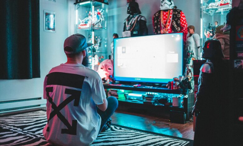 Back view of anonymous male sitting on floor near modern monitor and playing video game in room with comic stuff