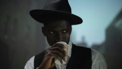 A stylish man in a fedora enjoys his coffee indoors, with a blurred cityscape backdrop.