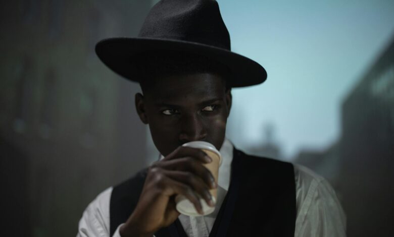 A stylish man in a fedora enjoys his coffee indoors, with a blurred cityscape backdrop.