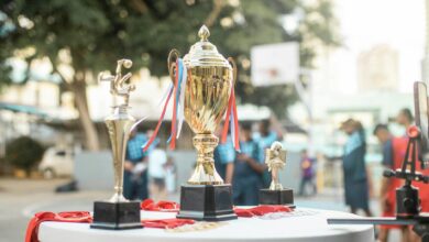 A golden trophy displayed on a table during an outdoor sports event with blurred players in the background.