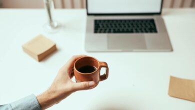 A person holds a brown coffee cup near a laptop on a clean white desk, perfect for remote work themes.