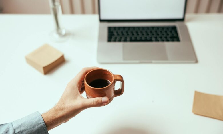 A person holds a brown coffee cup near a laptop on a clean white desk, perfect for remote work themes.