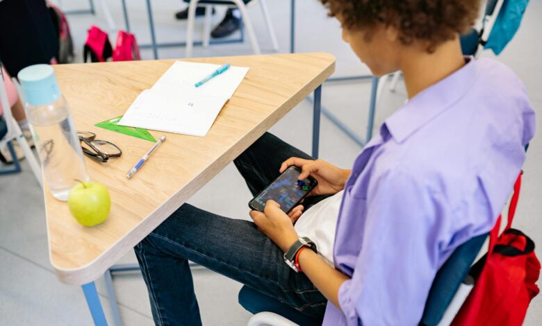 A child playing a mobile game at a school desk with school supplies nearby, captured indoors during class.