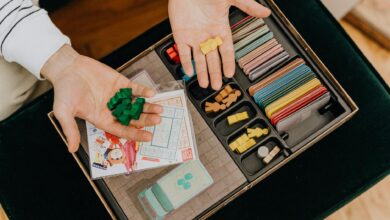 Colorful board game components being held in hands above a game box, top view.