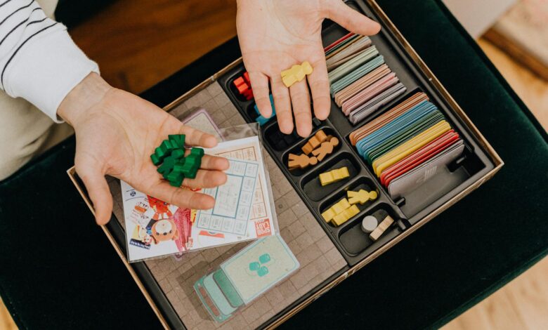 Colorful board game components being held in hands above a game box, top view.