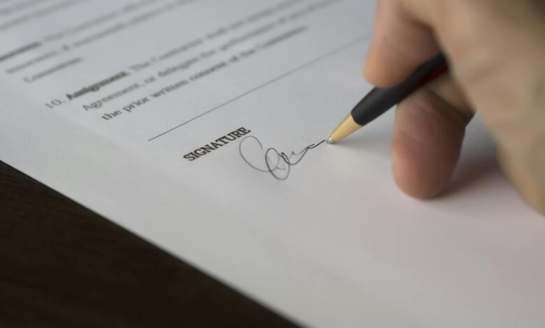 A hand signs a formal contract with a pen on a wooden desk.