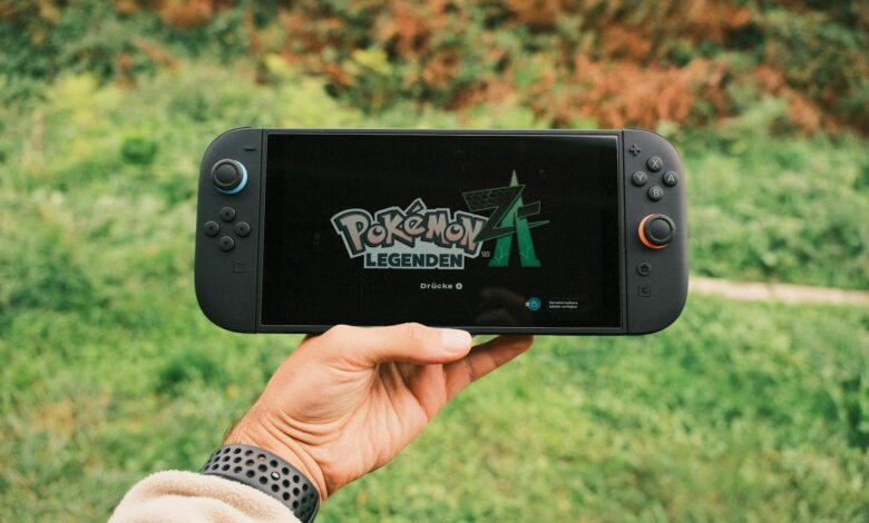A hand holding a handheld gaming console displaying the Pokémon Legends game screen outdoors.