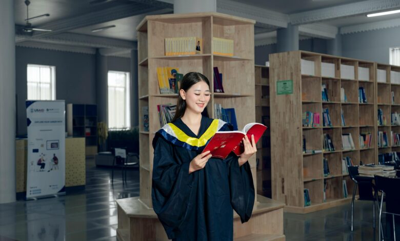 Woman in graduation gown reading a book in university library, symbolizing education and knowledge.