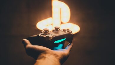 A hand holding a PlayStation controller with a blurred neon game symbol in the background.