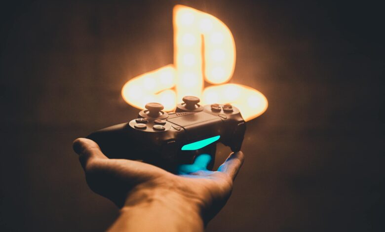 A hand holding a PlayStation controller with a blurred neon game symbol in the background.