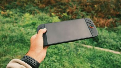 Close-up of a person's hand holding a black handheld gaming console against a green outdoor background.