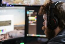 Side view concentrated bearded male in headphones using modern computer and looking at multiple monitors