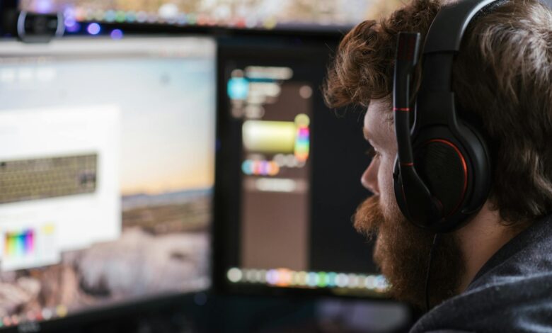 Side view concentrated bearded male in headphones using modern computer and looking at multiple monitors