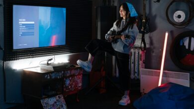 A young woman plays guitar in a stylish room with neon lighting and a wall-mounted screen.