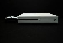 Close-up of a white gaming console with a black controller on a dark background.