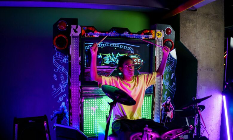 A young musician passionately plays electronic drums at an illuminated arcade setup.