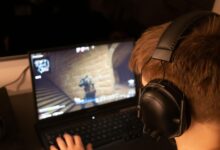 Teenage boy wearing headphones focused on playing a video game on a laptop screen indoors.