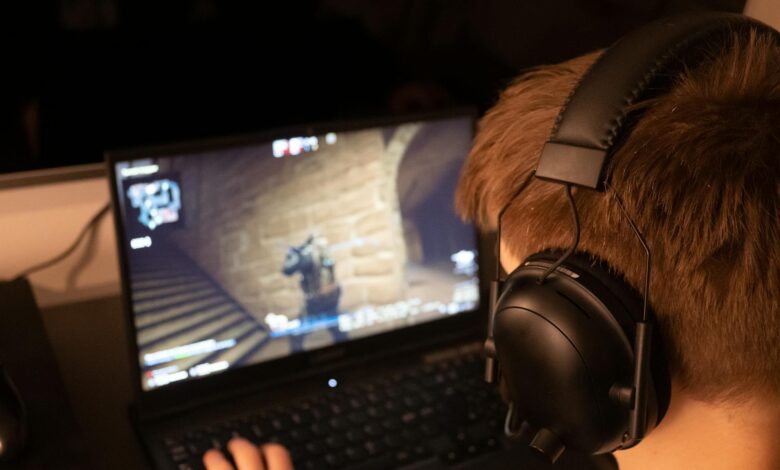 Teenage boy wearing headphones focused on playing a video game on a laptop screen indoors.