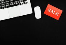 Minimalist flat lay of a laptop, mouse, and a red sale sign on black background.
