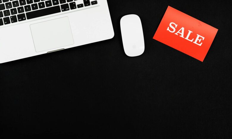 Minimalist flat lay of a laptop, mouse, and a red sale sign on black background.