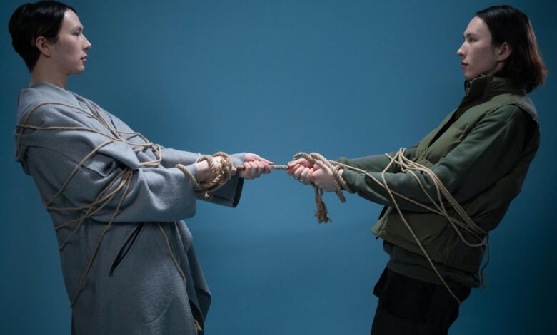 Two adults engaged in a symbolic tug of war, wrapped in rope against a blue background.