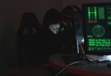 An anonymous hacker wearing a Guy Fawkes mask sits at a computer in a dimly lit room, engaged in cyber activities.