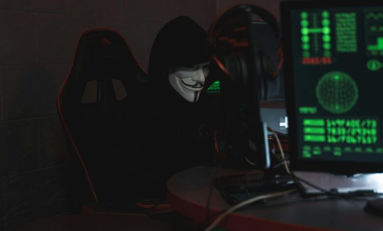 An anonymous hacker wearing a Guy Fawkes mask sits at a computer in a dimly lit room, engaged in cyber activities.