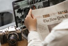 Close-up of a person reading The New York Times by a laptop and camera.