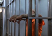 A tattooed inmate's arms rest on jail cell bars, wearing an orange uniform inside a prison.
