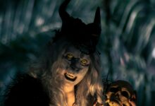A costumed figure with horns and yellow eyes holds a skull in a dark forest setting, creating a spooky vibe.